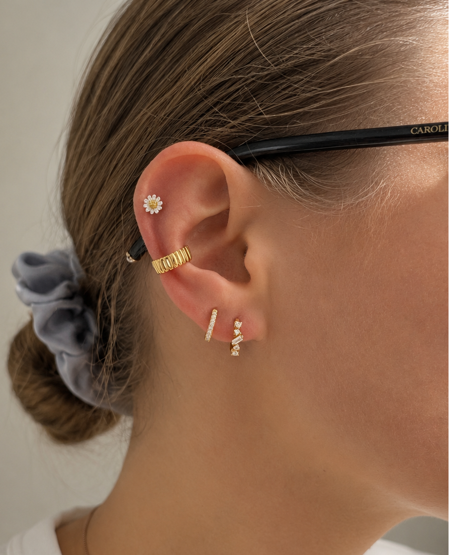 A close-up of a person's ear wearing multiple delicate gold and silver earrings, including a flower-shaped stud, a textured gold hoop, and small bar and dangle earrings. The person has brown hair tied back with a gray scrunchie and is wearing a white top.
