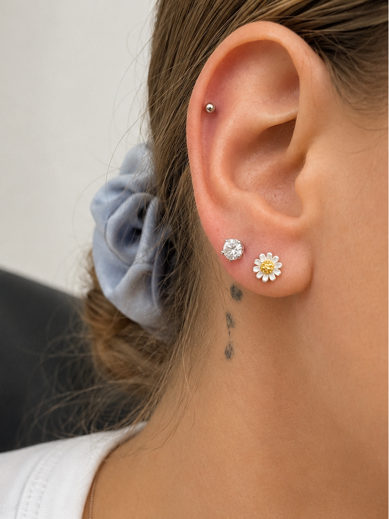 Close-up of a person's ear wearing two stud earrings: a small silver hoop piercing and a daisy-shaped stud with a silver center and white petals, shown against the neck and hair.