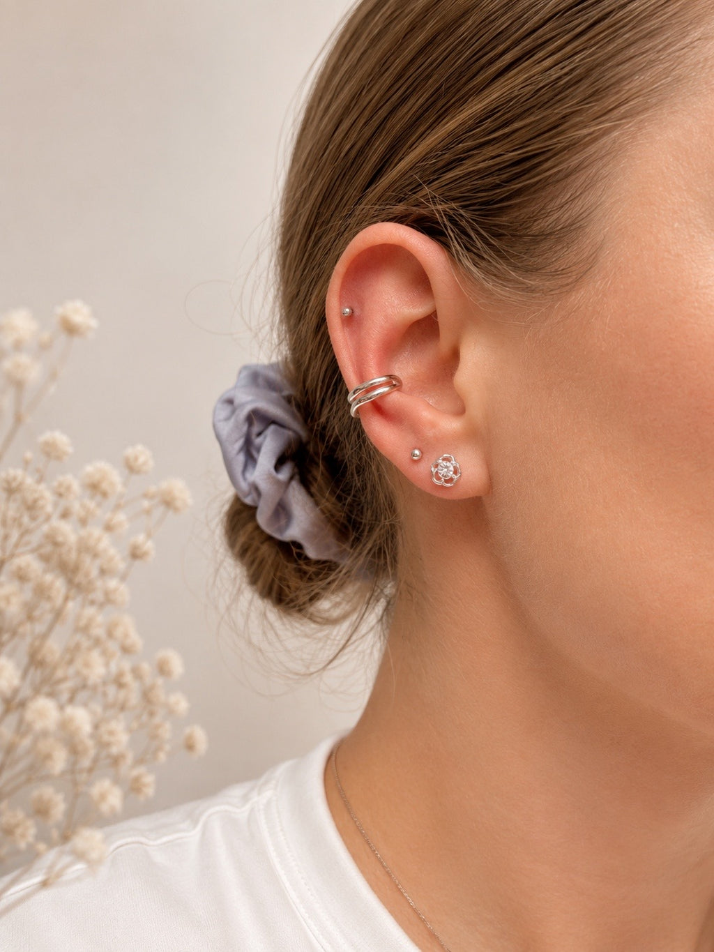 Close-up of a person's ear showcasing multiple silver-toned ear piercings, including a hoop in the cartilage, a small stud, and a cluster stud, with a lavender scrunchie in the hair.