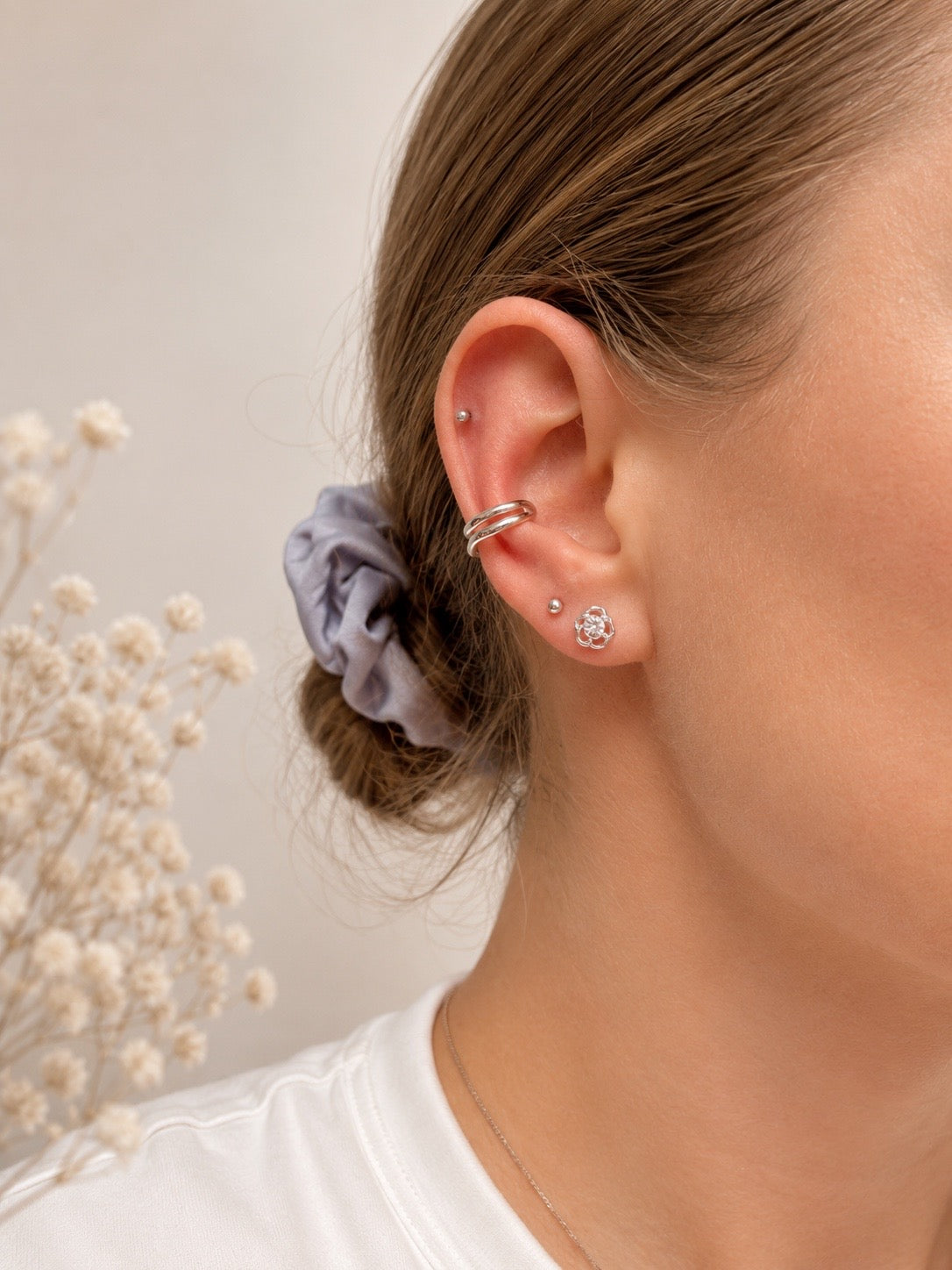 Close-up of a person's ear showcasing multiple silver-toned ear piercings, including a hoop in the cartilage, a small stud, and a cluster stud, with a lavender scrunchie in the hair.