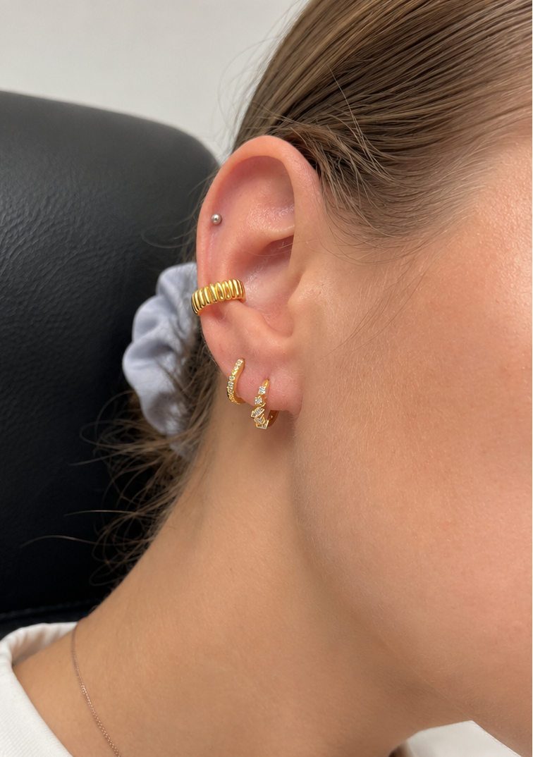 A close-up of a person's ear wearing multiple gold earrings, including a textured hoop on the lobe and stacked gold studs on the cartilage, with a light purple scrunchie in the hair.