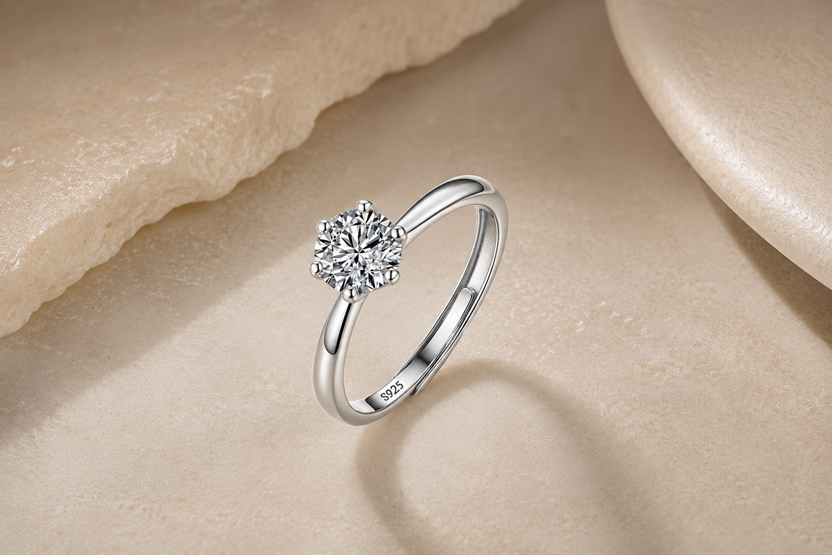 A silver solitaire engagement ring with a round brilliant-cut diamond, displayed on a textured beige stone surface.