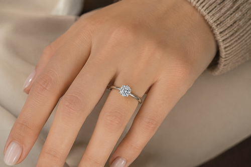 A close-up of a hand wearing a delicate silver solitaire ring with a round clear gemstone, set against a soft beige background.