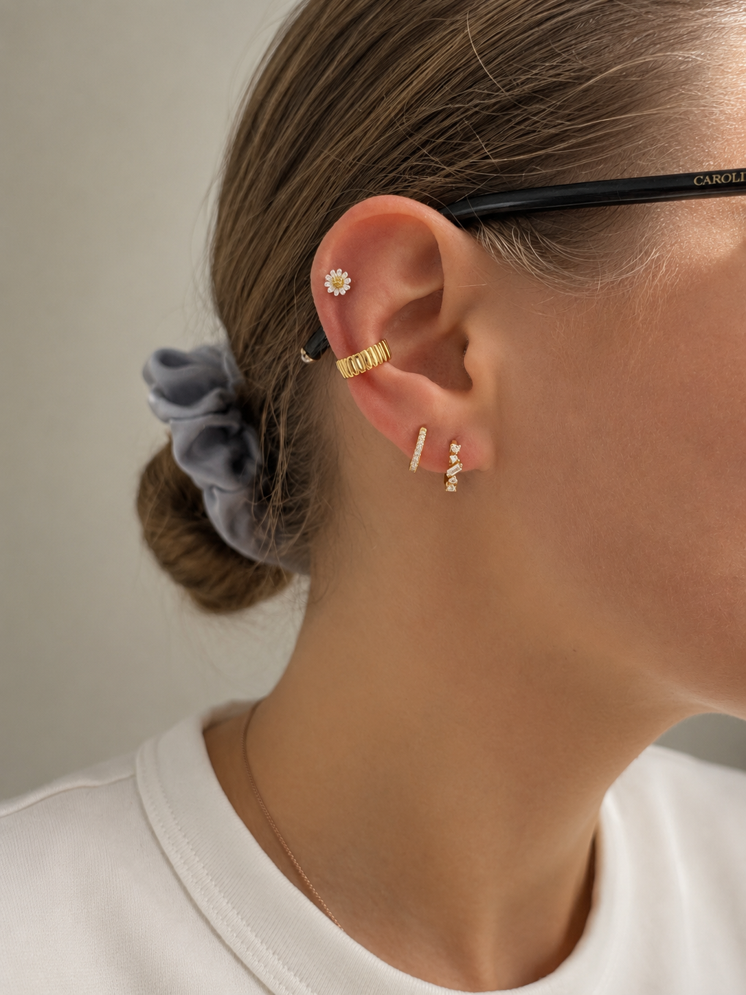 A close-up of a person's ear wearing multiple delicate gold and silver earrings, including a flower-shaped stud, a textured gold hoop, and small bar and dangle earrings. The person has brown hair tied back with a gray scrunchie and is wearing a white top.