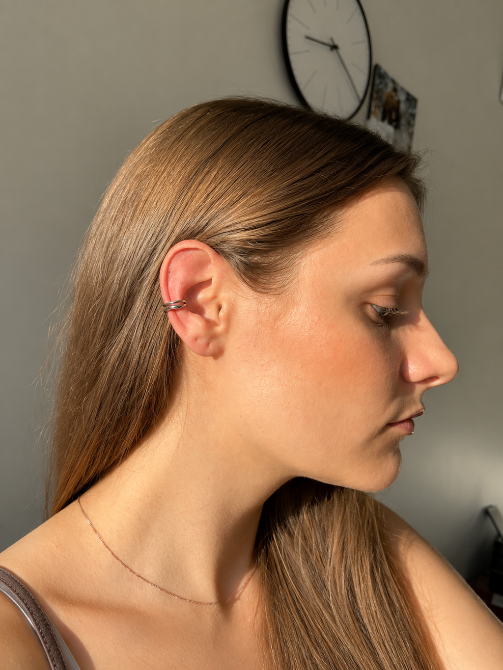 A side-profile portrait of a woman with long light brown hair wearing a silver hoop piercing in her cartilage ear, in warm natural light against a gray wall.