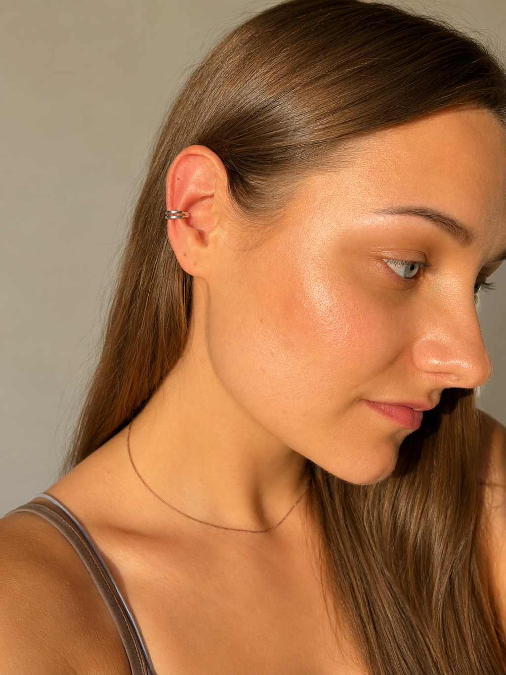 A close-up side profile of a woman with long brown hair wearing a delicate silver hoop piercing in her upper ear cartilage, along with a thin silver chain necklace, against a neutral gray background.