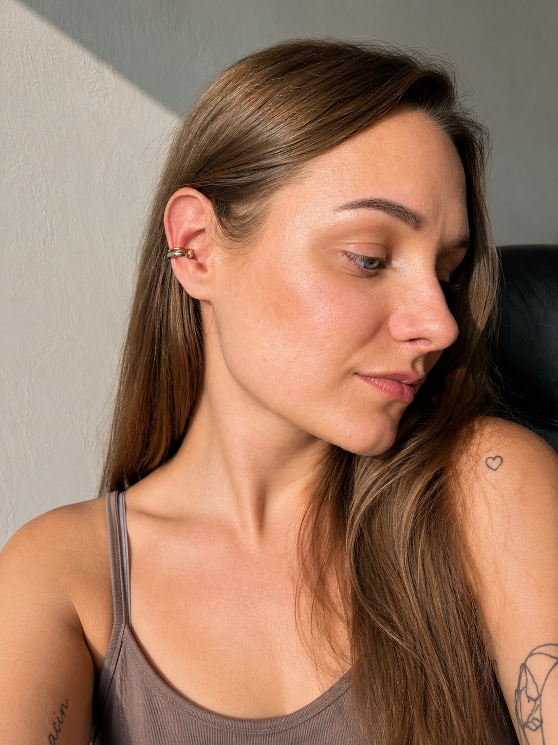 A woman with long brown hair wearing a silver-toned multi-prong ear cuff on her left ear, shown in a close-up portrait against a neutral gray wall.