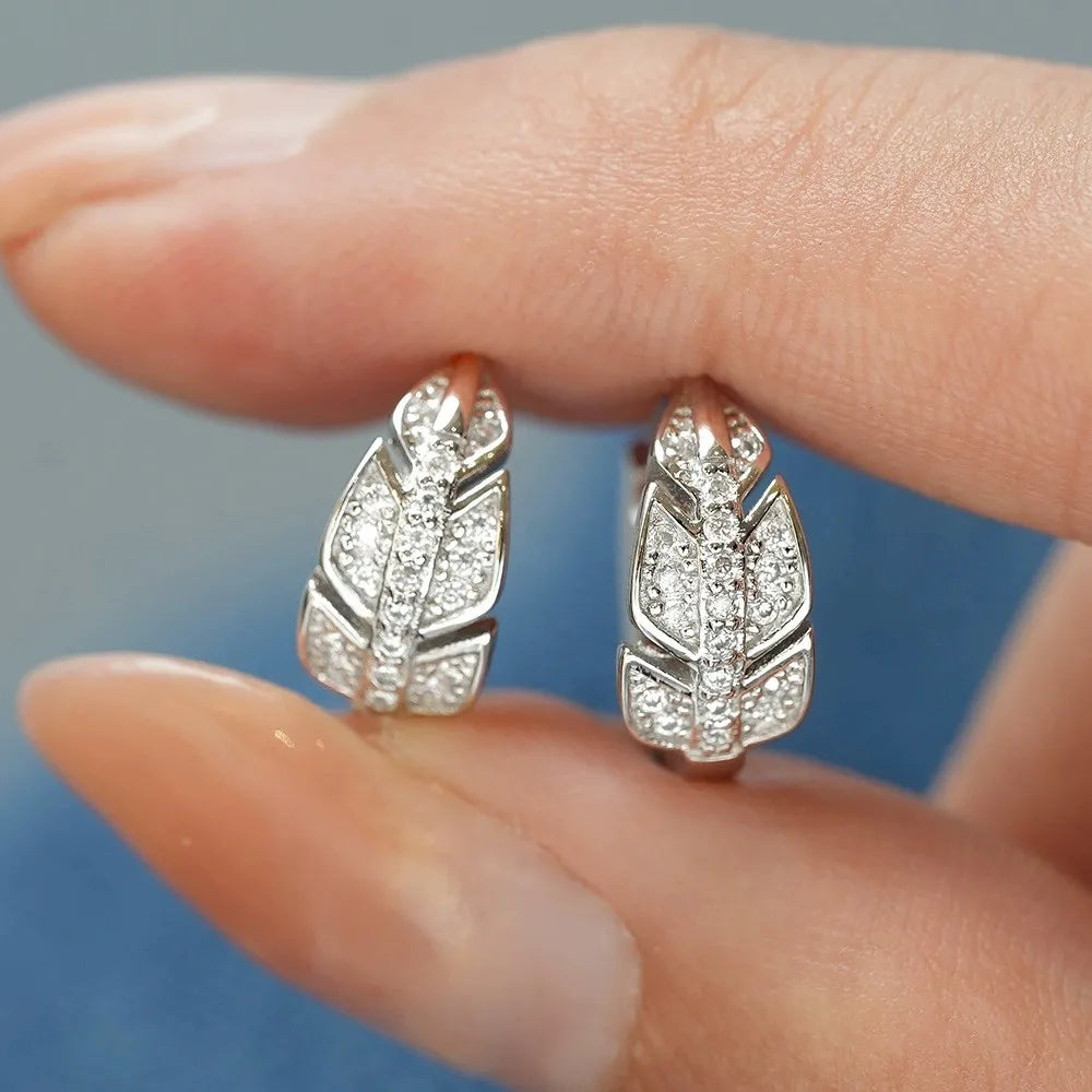 Silver feather-shaped earrings with gemstones held between fingers against a blurred background