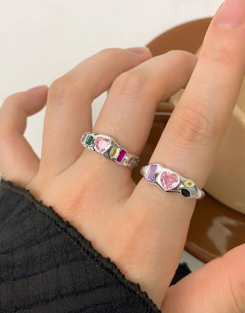 A close-up of a hand wearing two silver rings with colorful heart-shaped and rectangular gemstones, set against a soft neutral background.