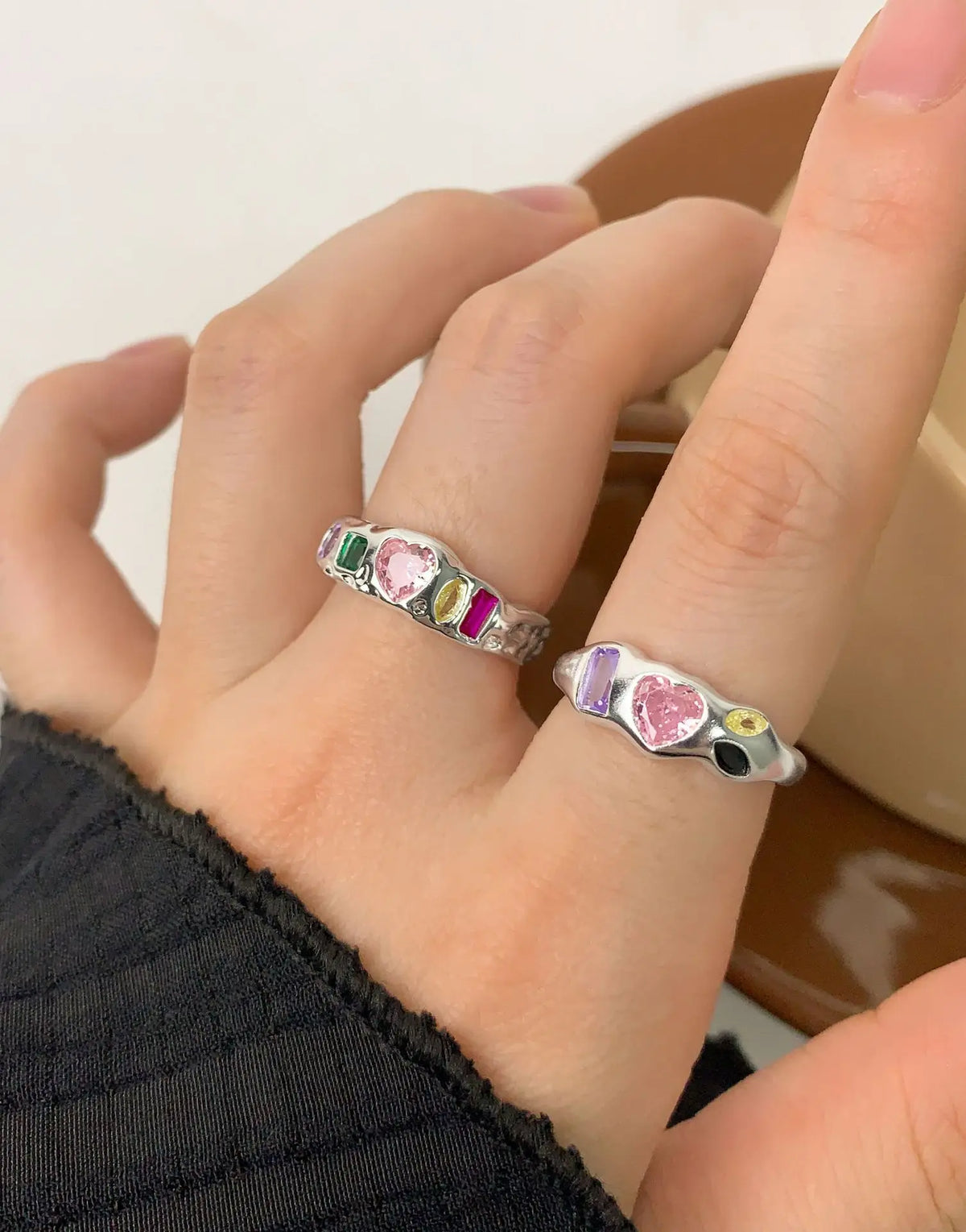 A close-up of a hand wearing two silver rings with colorful heart-shaped and rectangular gemstones, set against a soft neutral background.