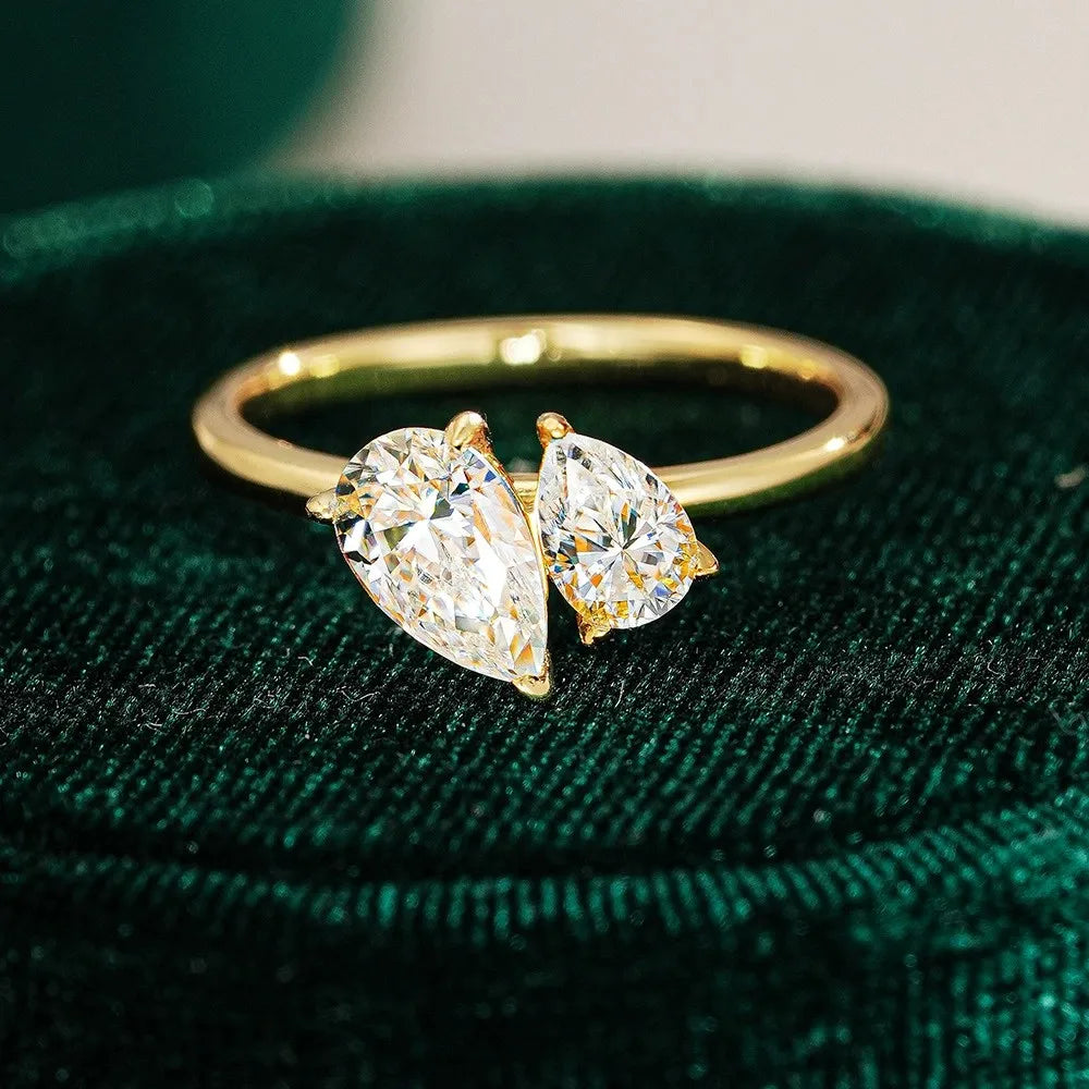 A gold ring with two pear-cut clear gemstones set in a split design, displayed on a dark green velvet surface.