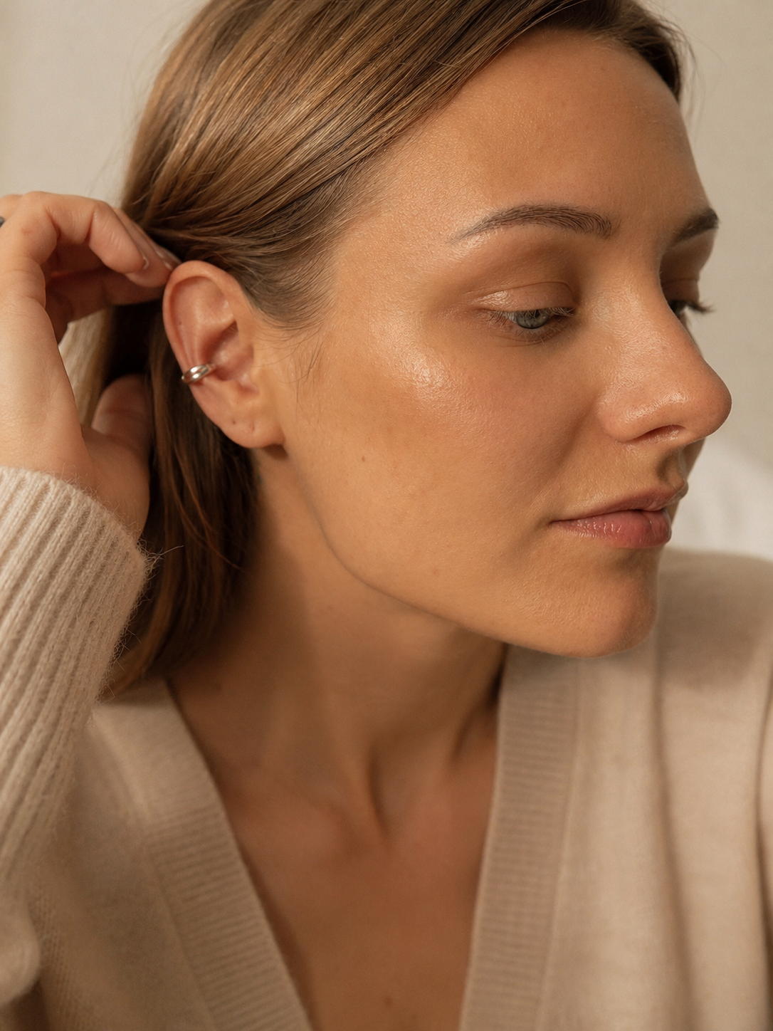 A close-up lifestyle shot of a woman wearing a delicate silver hoop earring in her ear, wearing a beige knit cardigan against a soft beige background.