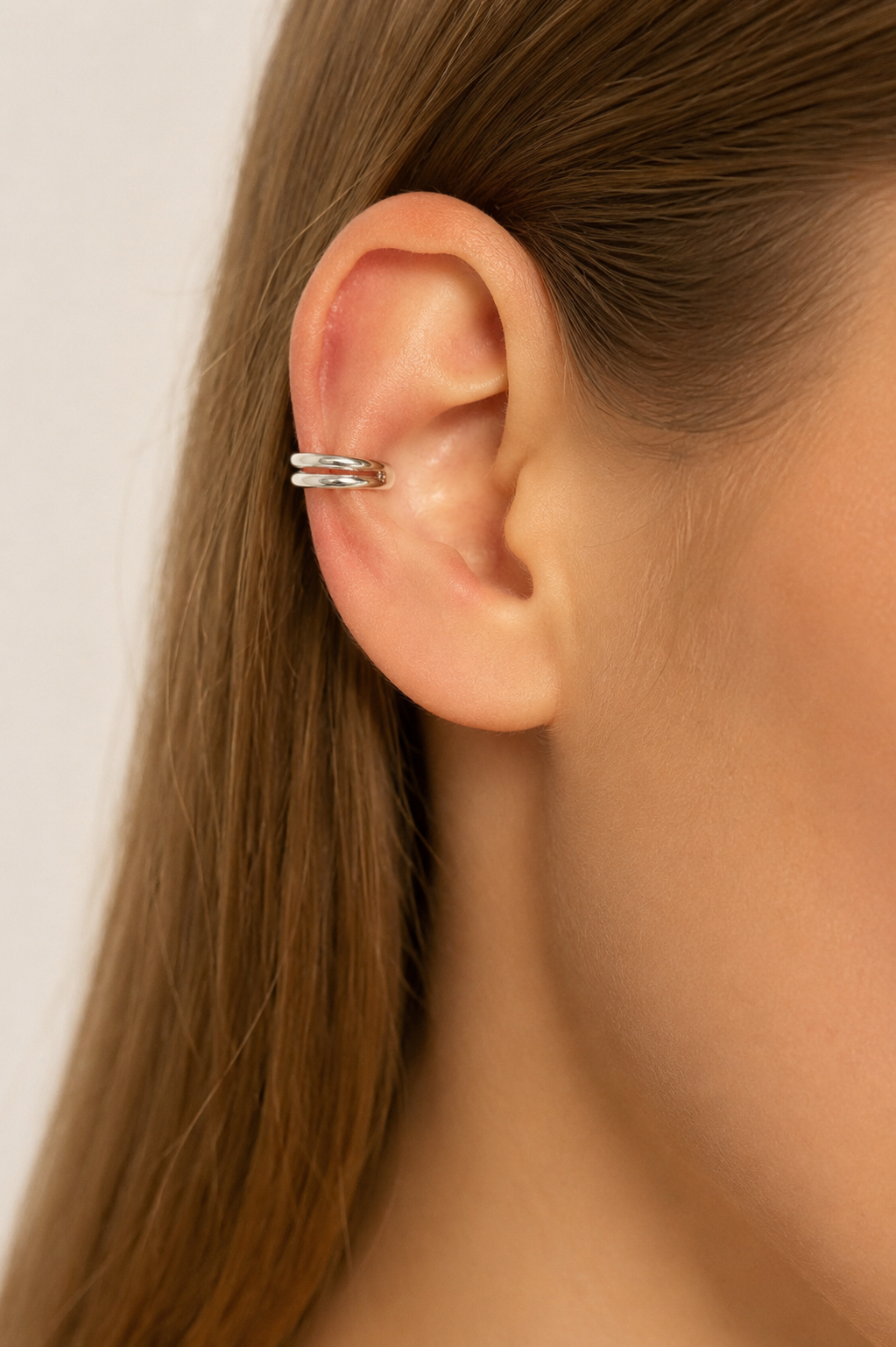 A close-up of a model's ear wearing a slim silver double-band ear cuff, with her hair pulled back and a plain light background.