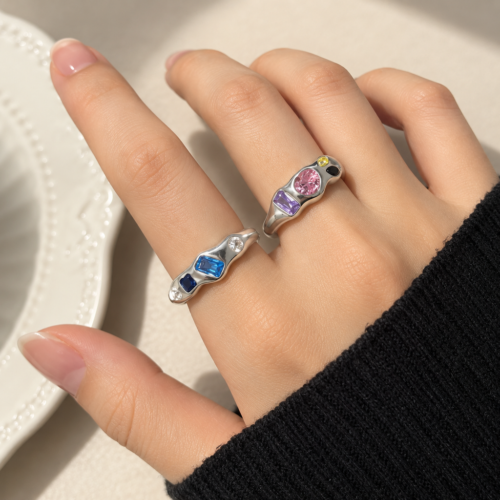 A close-up of a hand wearing two silver snake-shaped rings with colorful gemstones, one with a blue gemstone and the other with pink, purple, and yellow gemstones, set against a light background.