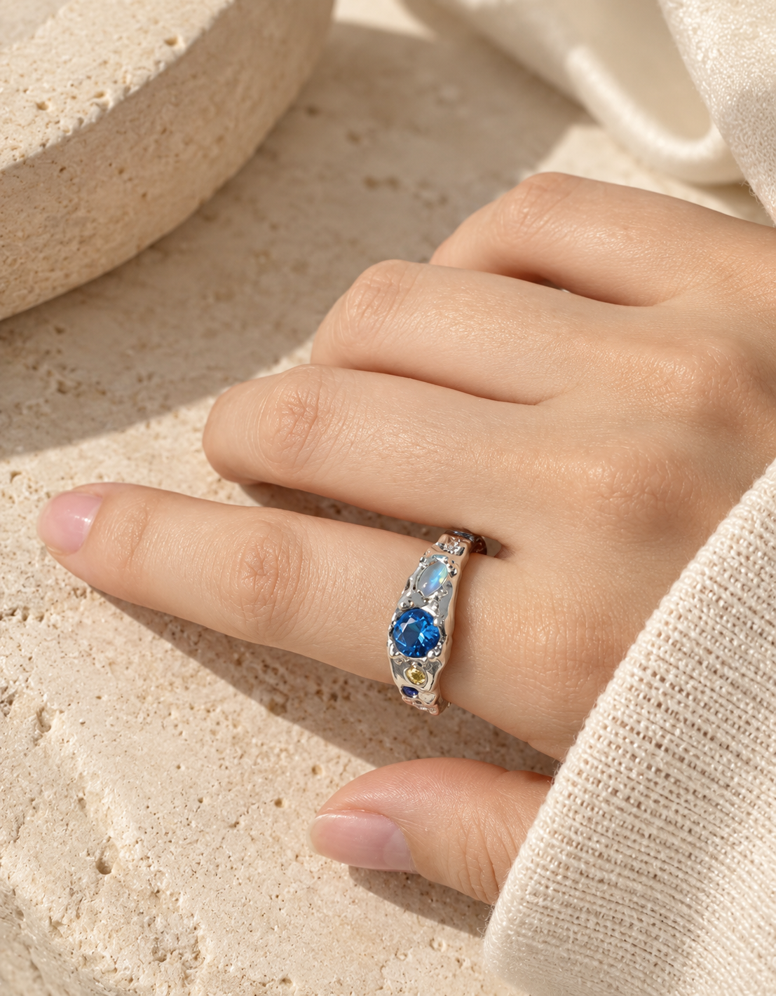 A close-up of a hand wearing a silver ring with a central round blue gemstone and smaller blue and clear stones along the band, set against a textured beige stone background.
