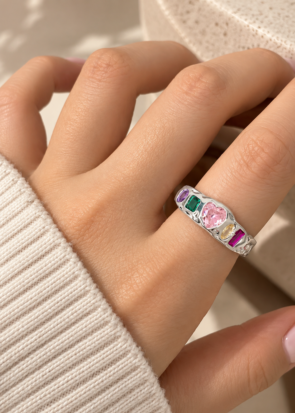 A close-up of a hand wearing a silver ring with a colorful heart and gemstone design, set against a soft beige background.