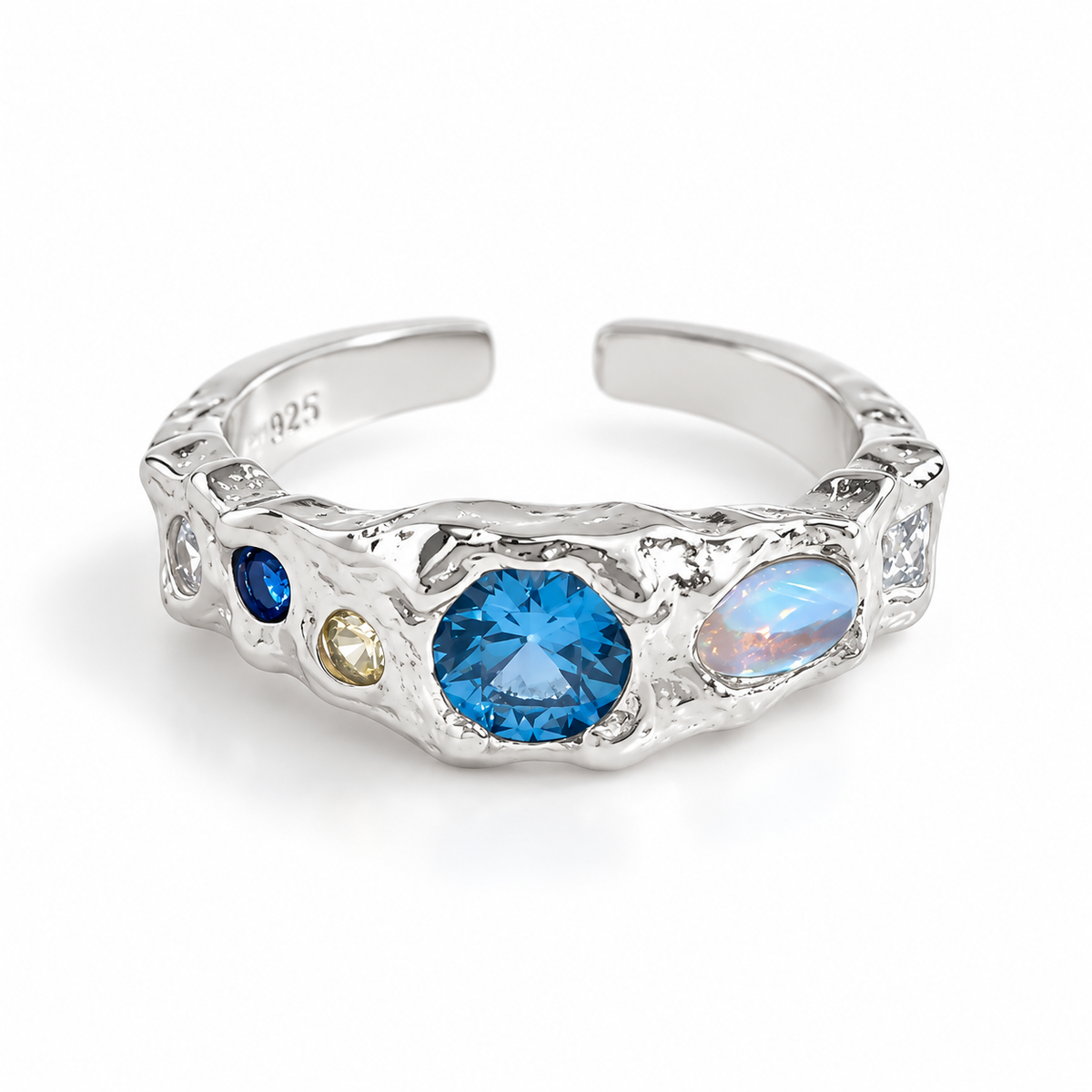 A silver-toned open ring band featuring a textured organic border and multiple inlaid gemstones, including a prominent round blue topaz, a larger iridescent blue opal, a smaller dark blue stone, a tiny yellow stone, and clear accent stones. The band is marked '925' on the inside.