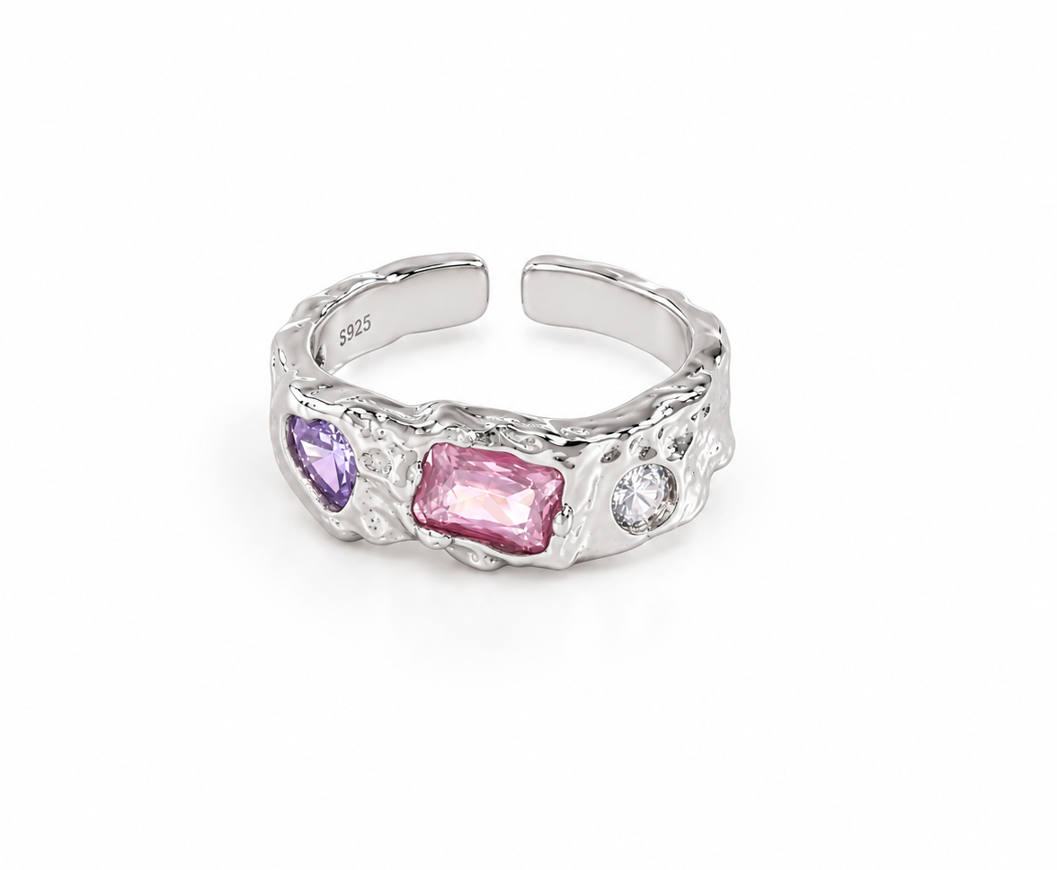 A silver adjustable ring featuring a central rectangular pink gemstone, a smaller oval purple gemstone on one side, and a small clear round gemstone on the other, set against a plain white background.