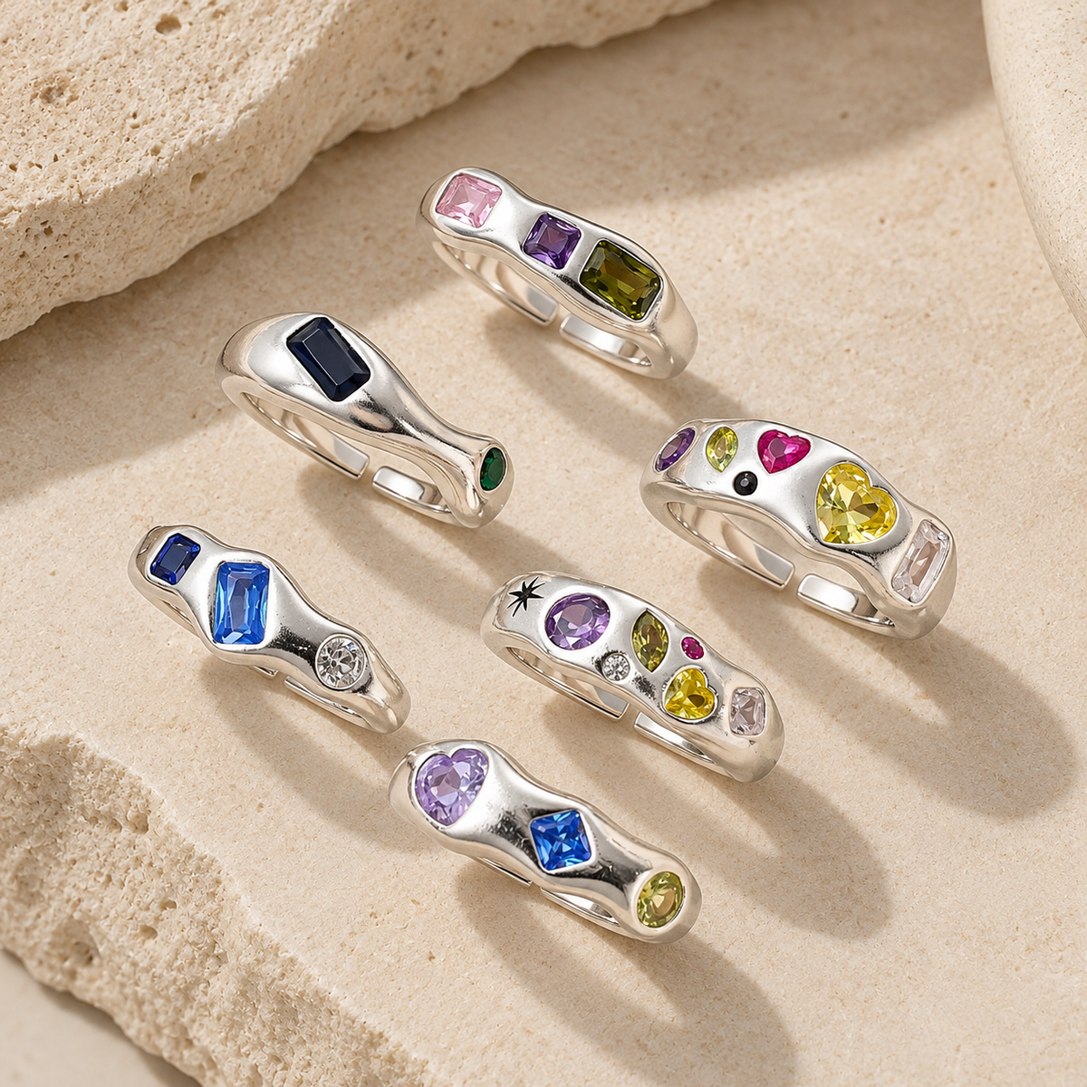 Seven silver rings with colorful gemstones are arranged on a textured beige stone surface.